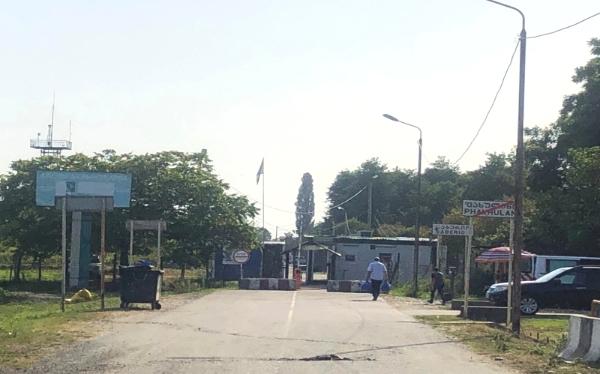 Border crossing checkpoint with a road leading to it, cars, trash container and man carrying a bag.